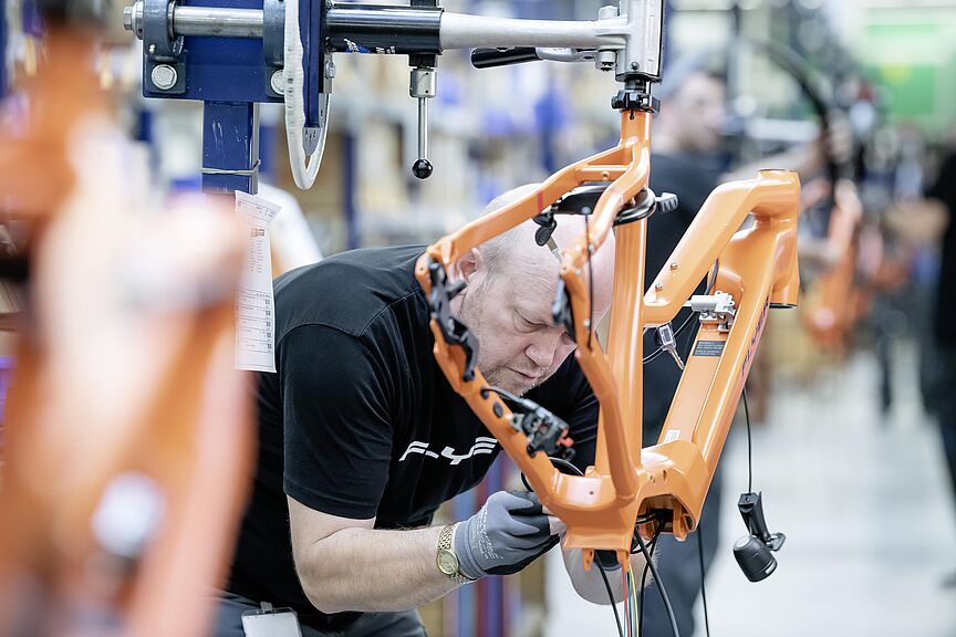 Elektrofahrrad-Montage in der Produktion Ein Mitarbeitender montiert den orangefarbenen Rahmen eines Elektrofahrrads in einer Fahrradmanufaktur.